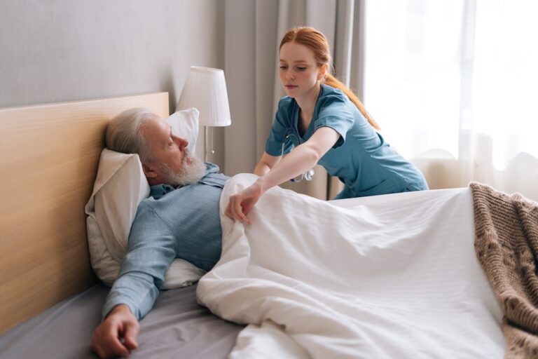 Side view of professional young female nurse helping senior adult male patient to lying on bed and covering him blanket at home.